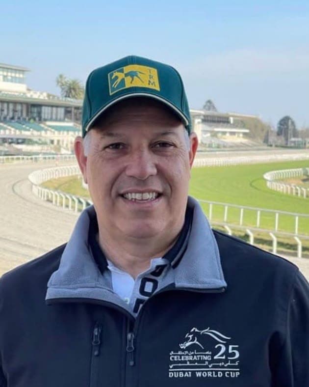 Antonio Cintra in a green cap and navy jacket, smiling in front of a racecourse grandstand