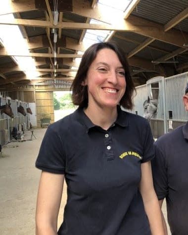 Audrey de Clerck smiling in a navy polo shirt inside a stable, horses in their stalls behind her