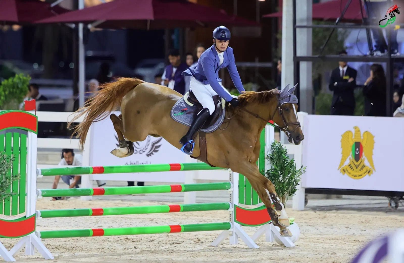 A rider in blue jumps a green and red fence in front of a banner with the Libyan emblem