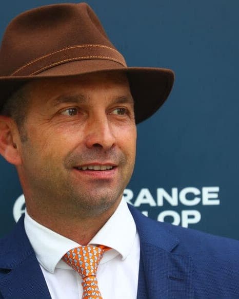 Damien de Watrigant in a brown fedora and navy suit in front of a Chantilly racecourse sign