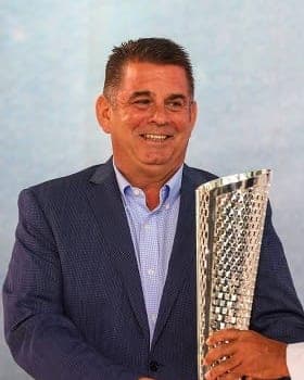 Doug Watson in a navy jacket and light blue shirt, smiling as he holds a silver trophy
