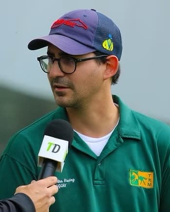 Julio Olascoaga in glasses and a navy cap, wearing a green polo and speaking into a microphone at the races