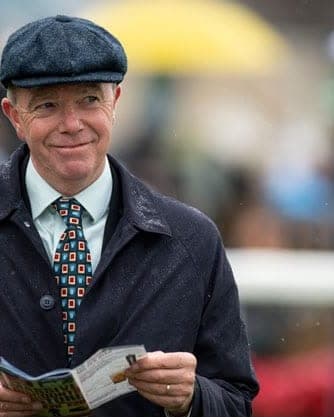 Karl Burke in a flat cap and dark coat, holding a racecard in the rain
