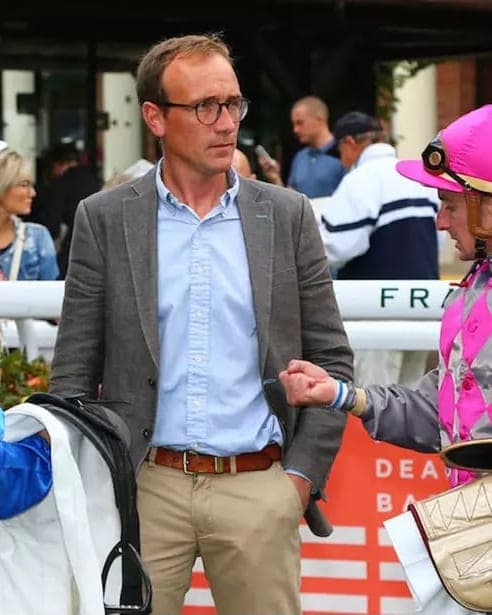 Xavier Thomas-Demeaulte in a grey blazer and glasses, greeting a jockey in pink and grey silks at the racecourse